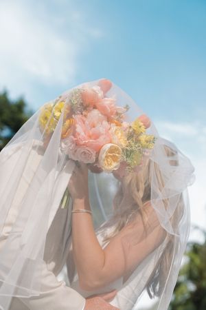 A flowing white wedding veil adorned with coral and yellow flowers blows softly in the breeze against a blue sky backdrop.