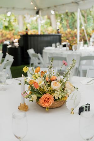 Elegant wedding table centerpiece with peach roses and mixed flowers in woven basket on white tablecloth under tent canopy.
