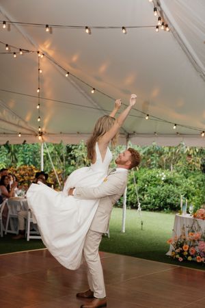 A romantic dance under a tent with string lights, featuring a flowing white dress and beige suit on a wooden dance floor.