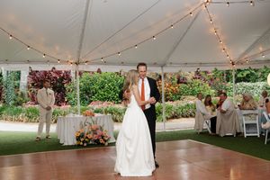 A romantic first dance takes place under a white tent with string lights and garden views during an outdoor wedding reception.