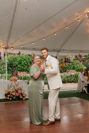 A dance moment under a white tent with string lights at a wedding reception features a sage green gown and beige suit.