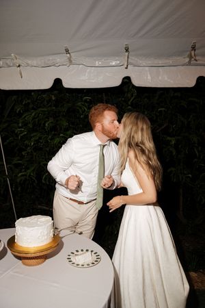 A couple in wedding attire shares a sweet moment while cutting their wedding cake under a white tent.