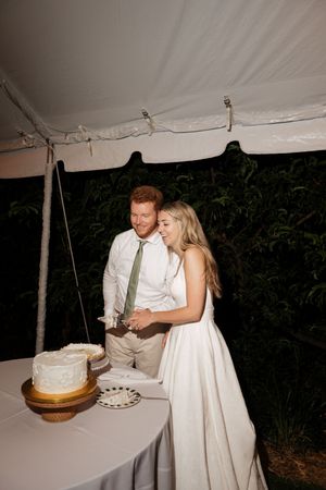 A couple in wedding attire cuts their white wedding cake together under a reception tent at night.