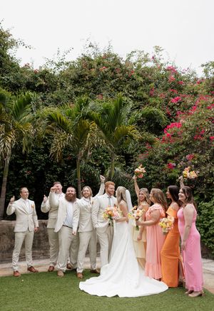 Wedding party in cream colored suits and colorful dresses poses outdoors with tropical plants and pink flowers in background.