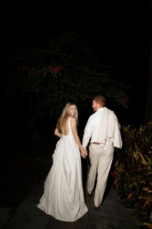 A couple in elegant wedding attire walks away into a dark garden at night, with the bride's white dress trailing behind.