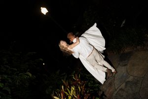 A couple in white formal attire embrace in a romantic nighttime garden scene illuminated by outdoor lighting.