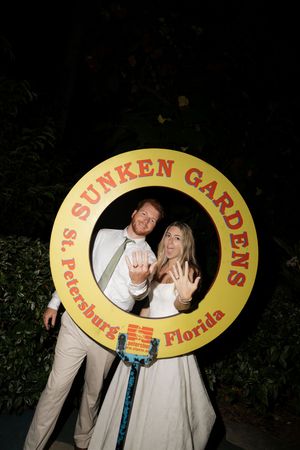 Couple poses with rings behind a yellow Sunken Gardens sign frame in St. Petersburg, Florida at night.