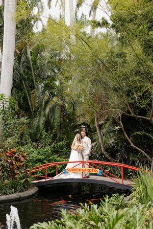 A tropical garden setting features a red footbridge over a pond surrounded by lush palm trees and greenery.