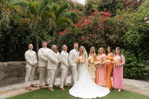 Outdoor wedding party poses in peach and tan attire against lush tropical garden backdrop with flowering bougainvillea.