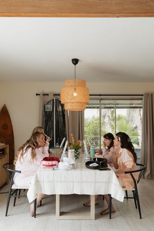 Friends gather around a white dining table under a woven pendant light while enjoying food and conversation at home.