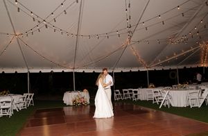 A couple shares a romantic dance under twinkling string lights at an outdoor evening reception with white folding chairs.