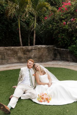 A couple in wedding attire relaxes on green grass with tropical flowers and palm trees in the garden background.