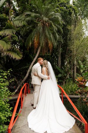 A couple embraces on a garden bridge beneath tropical palm trees and lush greenery during their wedding ceremony.