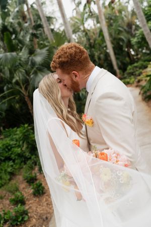 Romantic wedding couple shares a tender kiss in a lush tropical garden setting with palm trees and peach flowers.