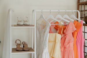 Pastel dresses hanging on white hangers next to a shelf displaying beige sandals against a white wall.