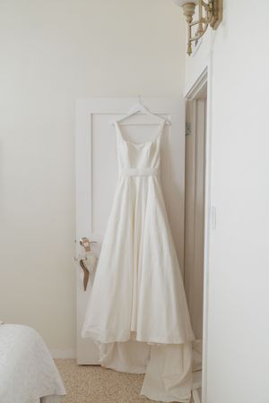 A classic white wedding dress with a long train hangs elegantly on a hanger against a light-colored wall.
