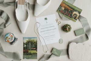 Wedding details flatlay with invitation, mint boxes, vintage postcards, gold rings, and white shoes on light gray fabric.