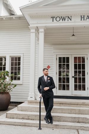 A groom in a black suit poses on the white steps of a historic Town Hall building during a formal occasion.