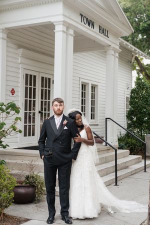 A couple poses in formal wedding attire on the steps of a white Town Hall building with classic columns.