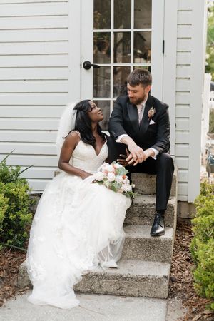 A couple sits together on church steps in wedding attire while holding a pink bouquet.