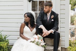A couple sits together on stone steps outside a white house, with the bride holding a pink and white bouquet.
