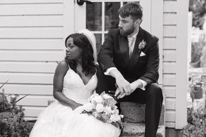 Black and white wedding photo of newlyweds sitting together outside a white building with rustic siding.