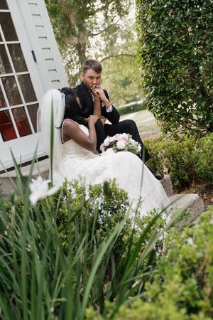 A person in formal attire sits thoughtfully on white steps in a garden setting surrounded by greenery and flowers.