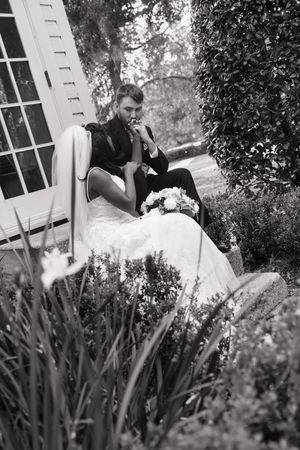 Black and white artistic photo showing a person in dark clothing sitting pensively in a garden setting near a white wall.