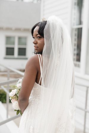 A bride in a flowing white wedding gown and veil holds a bouquet as she poses in profile against white architecture.