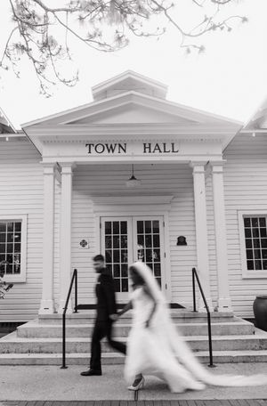 Black and white photo showing movement of two people walking past a traditional white clapboard New England town hall entrance.