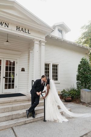 A wedding couple shares a romantic moment on the steps of a white Town Hall building with classic colonial architecture.