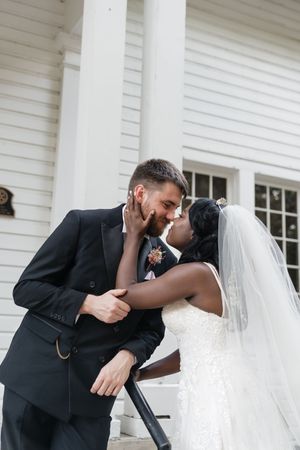 A bride in a white dress and groom in a dark suit share a romantic kiss on steps of a white columned building.