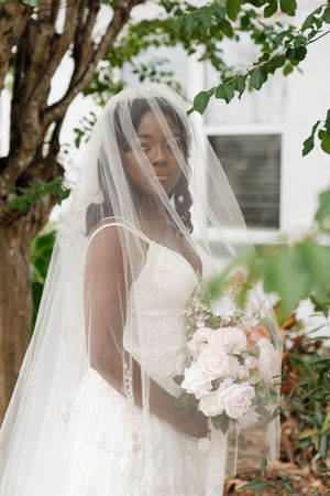 A bride in a white lace wedding dress and flowing veil poses elegantly among green trees and foliage.