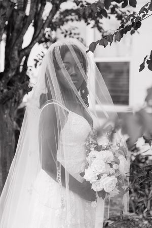Black and white photo of a bride in elegant lace wedding dress with long veil holding white rose bouquet in garden setting.