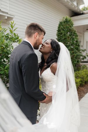 A couple shares an intimate moment on their wedding day in front of a white-painted brick wall with greenery.
