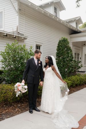 A couple in wedding attire stands together on a pathway outside a white church building with landscaping.