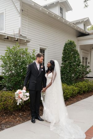 A bride and groom share a loving moment outside a white chapel building with beautiful landscaping and a bridal bouquet.
