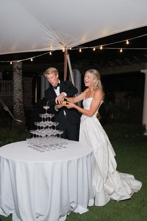 Bride and groom pouring champagne over an elegant glass tower at outdoor wedding reception.