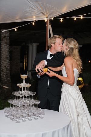 Bride and groom kissing while pouring champagne tower at outdoor wedding reception under string lights.