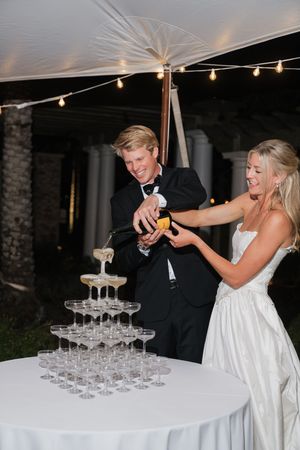 Smiling newlyweds pouring champagne over an elegant tiered champagne tower at their outdoor wedding reception at night.