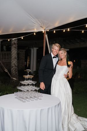 Bride and groom smiling beside a champagne tower at their elegant outdoor wedding reception under a white tent.