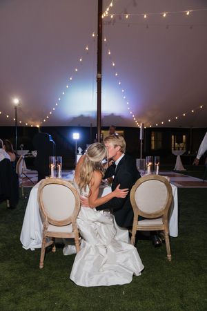 Bride and groom share a romantic kiss at sweetheart table under glowing string lights in wedding tent.
