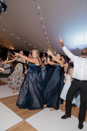 Wedding guests in navy gowns dancing energetically under string lights on white dance floor.