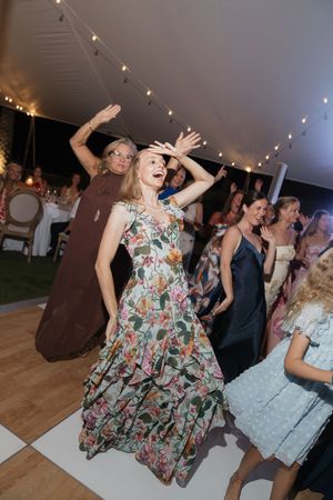 Guest in floral dress dances joyfully at an outdoor wedding reception under string lights in a white tent.