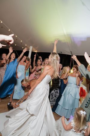 Bride dancing energetically with guests at a lively wedding reception under a white tent with string lights.