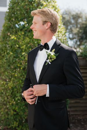 Smiling groom in black tuxedo with floral boutonniere standing outdoors on wedding day.