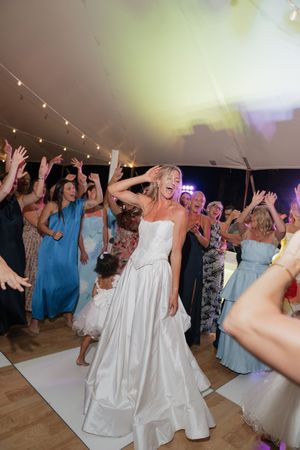 Bride in white gown dancing joyfully with guests at wedding reception under string lights in a tent.