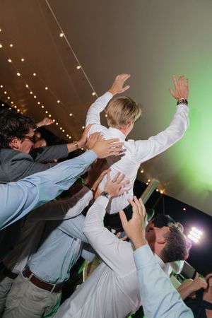 Groomsmen lifting the groom into the air during a lively wedding reception celebration under string lights.