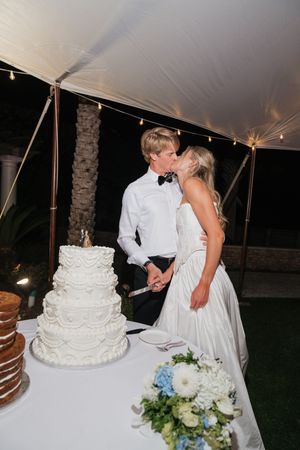 Bride and groom share a kiss while cutting their white four-tier wedding cake at an outdoor evening reception.