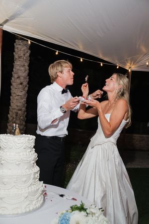 Smiling bride and groom feeding each other wedding cake at their outdoor nighttime reception.